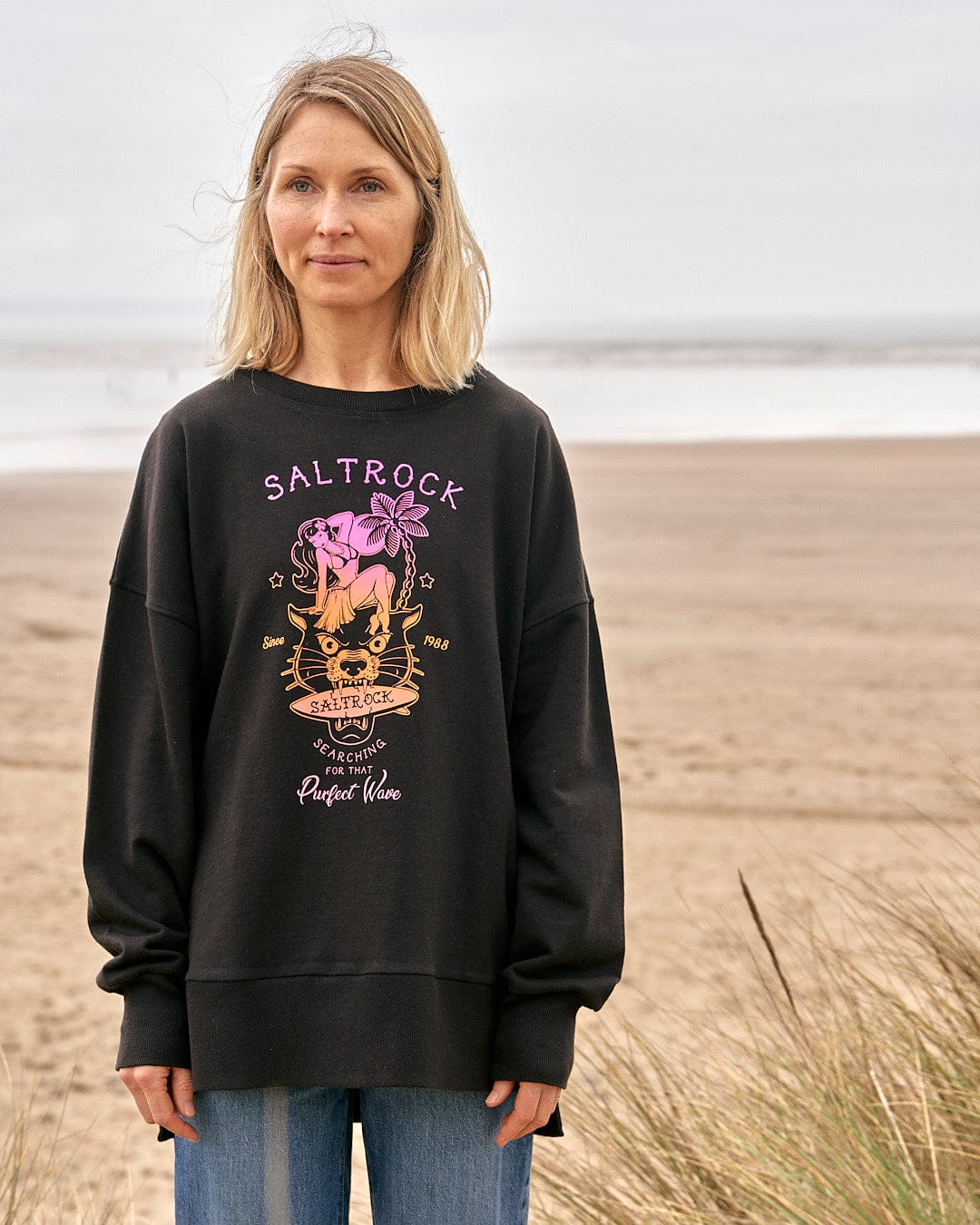 A woman wearing a Saltrock Purfect Wave Gradient - Womens Boyfriend Fit Sweat - Black sweatshirt standing on the beach.