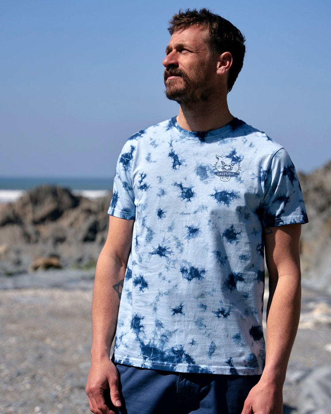 A man is standing on the beach wearing a Saltrock - Purfect Wave Mens Tie Dye Short Sleeve T-shirt in Light Blue.