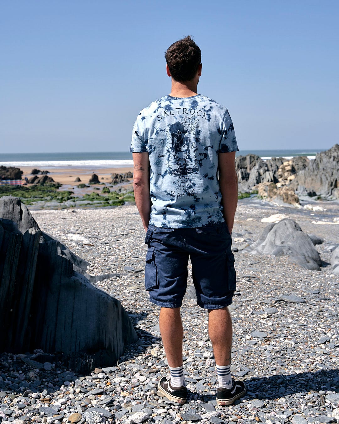A man standing on a beach looking at the ocean wearing a Saltrock Purfect Wave - Mens Tie Dye Short Sleeve T-shirt in Light Blue.