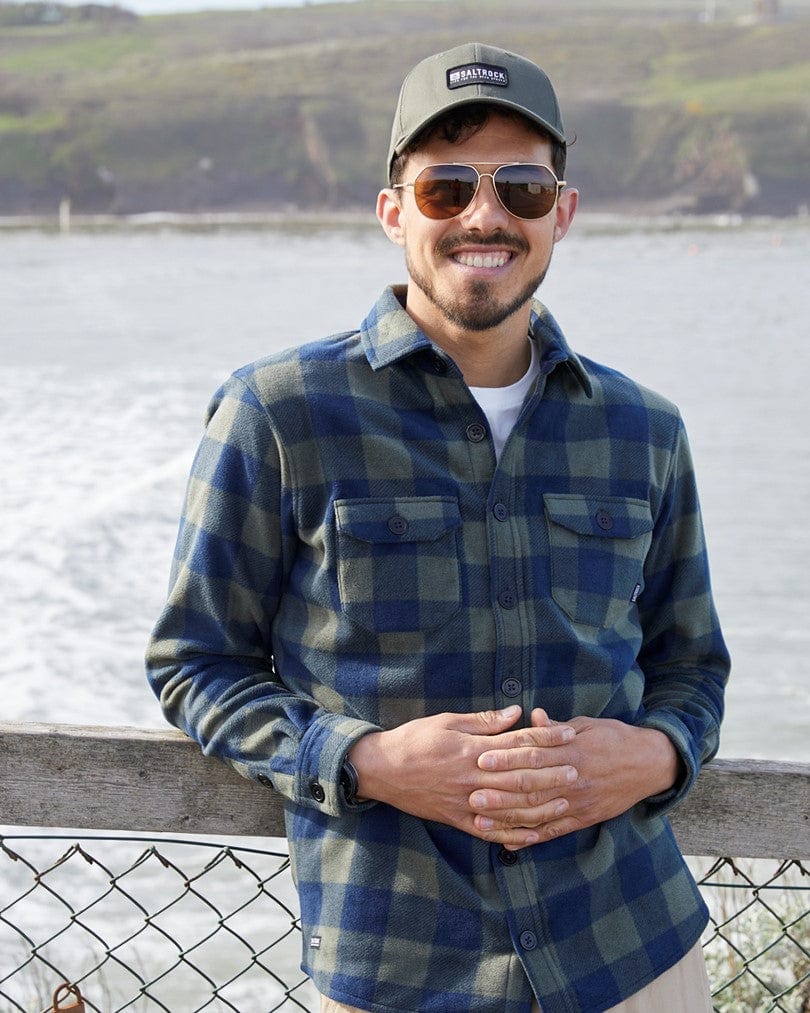 A man wearing a Saltrock Waldron - Mens Fleece Shirt in Dark Green and sunglasses smiles while standing by a railing with a blurred coastal background.