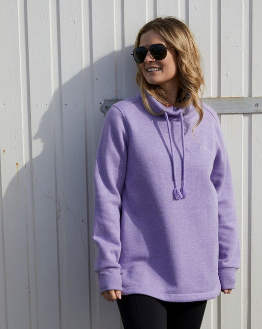 Woman in sunglasses standing and smiling, wearing a Saltrock Harper - Womens Longline Pop Sweat - Lilac against a corrugated metal wall.