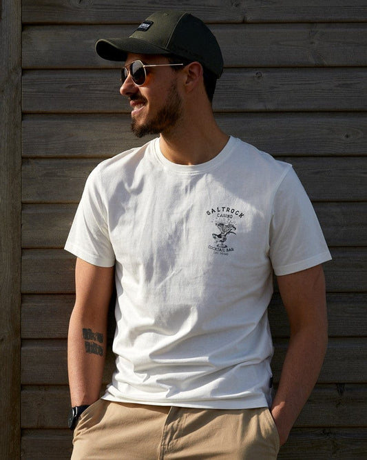 A man in a Saltrock Vegas Cocktail - Mens Short Sleeve T-Shirt - White and black cap smiles and looks to the side, leaning against a wooden wall, with a visible tattoo on his right arm.