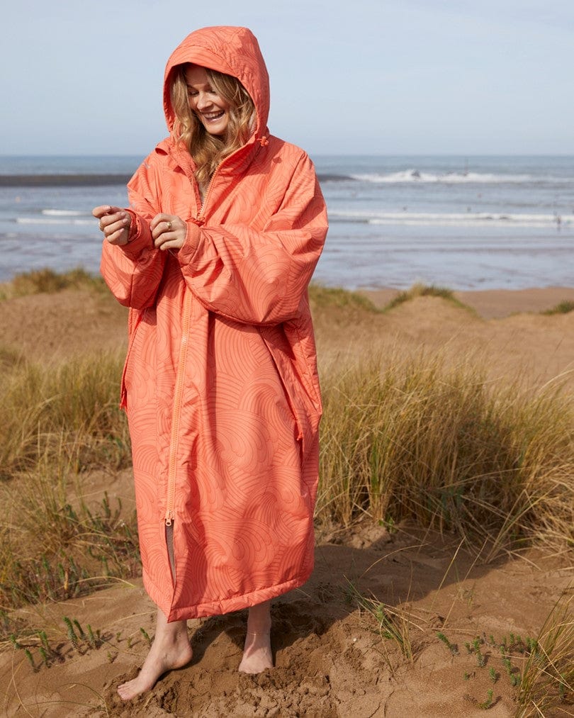 Woman in a Saltrock Recycled Four Seasons Changing Robe - Light Orange, smiling and standing on a sandy beach with grassy dunes and the ocean in the background.