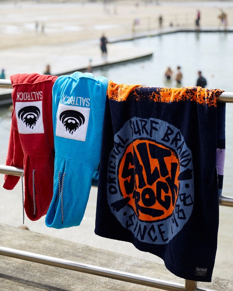 Three Saltrock Corp Changing Towels - Blue hanging on a railing with a beach and swimmers in the background.