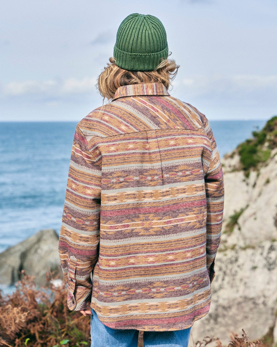 A man is standing on a rocky cliff wearing Saltrock's Quinn - Womens Heavy Overshirt - Brown, looking at the ocean.