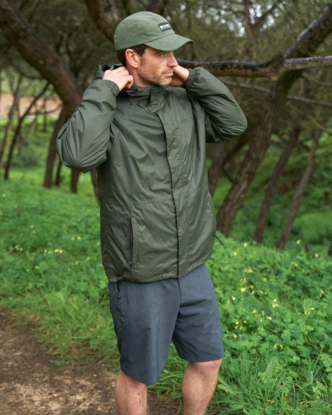 Man in a forest adjusting the hood of his Saltrock Rainier - Mens Packable Waterproof Jacket - Green, wearing a cap and shorts, with lush greenery and trees in the background.