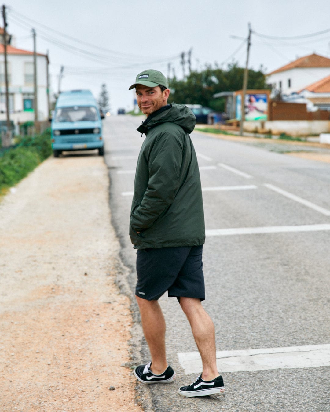 Man in a Saltrock Rainier - Mens Packable Waterproof Jacket in Green and black shorts smiling over his shoulder while standing on a suburban road.