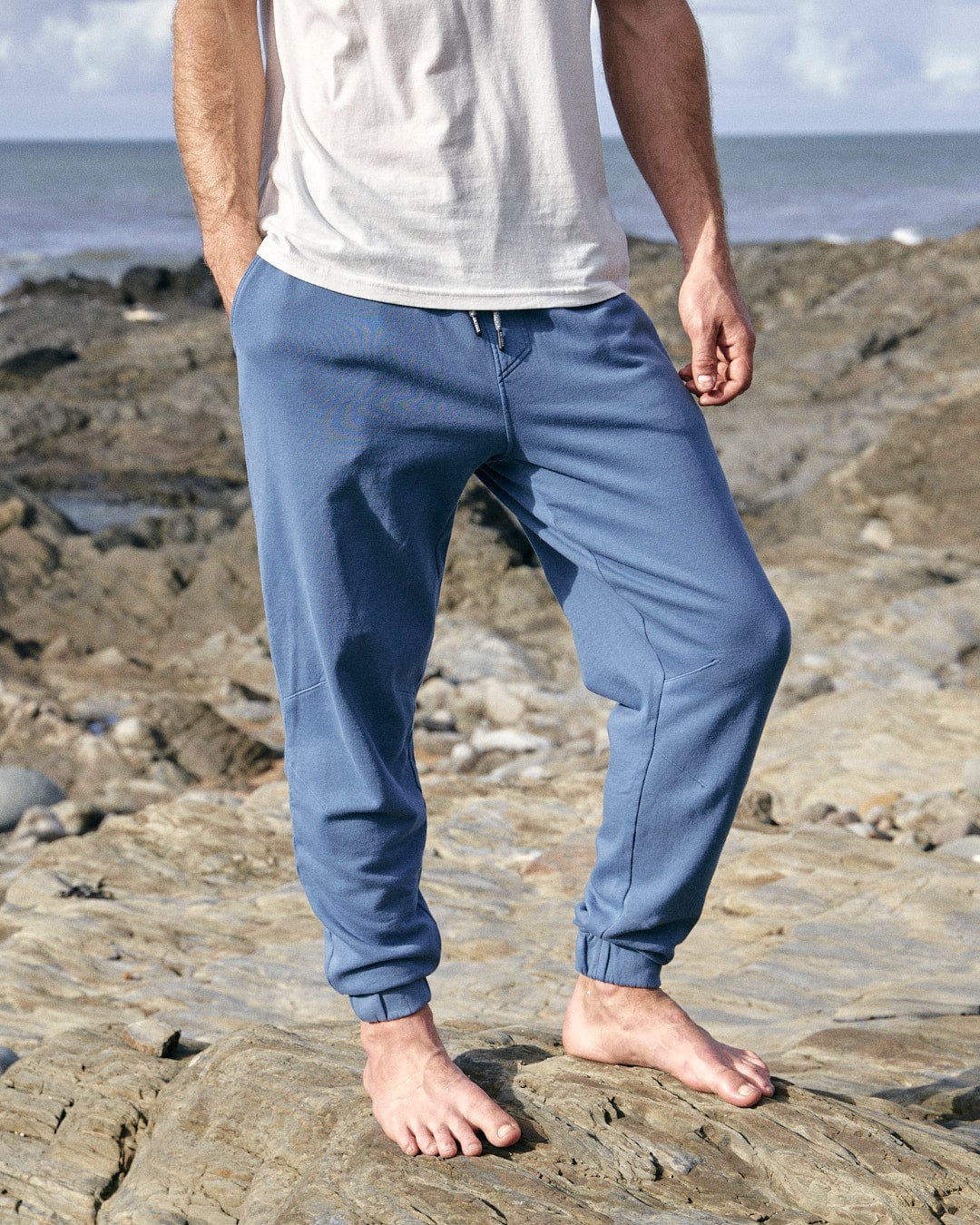 A man standing on rocks next to the ocean wearing Saltrock's Ralfen - Mens Slimfit Sweatpants in Blue.