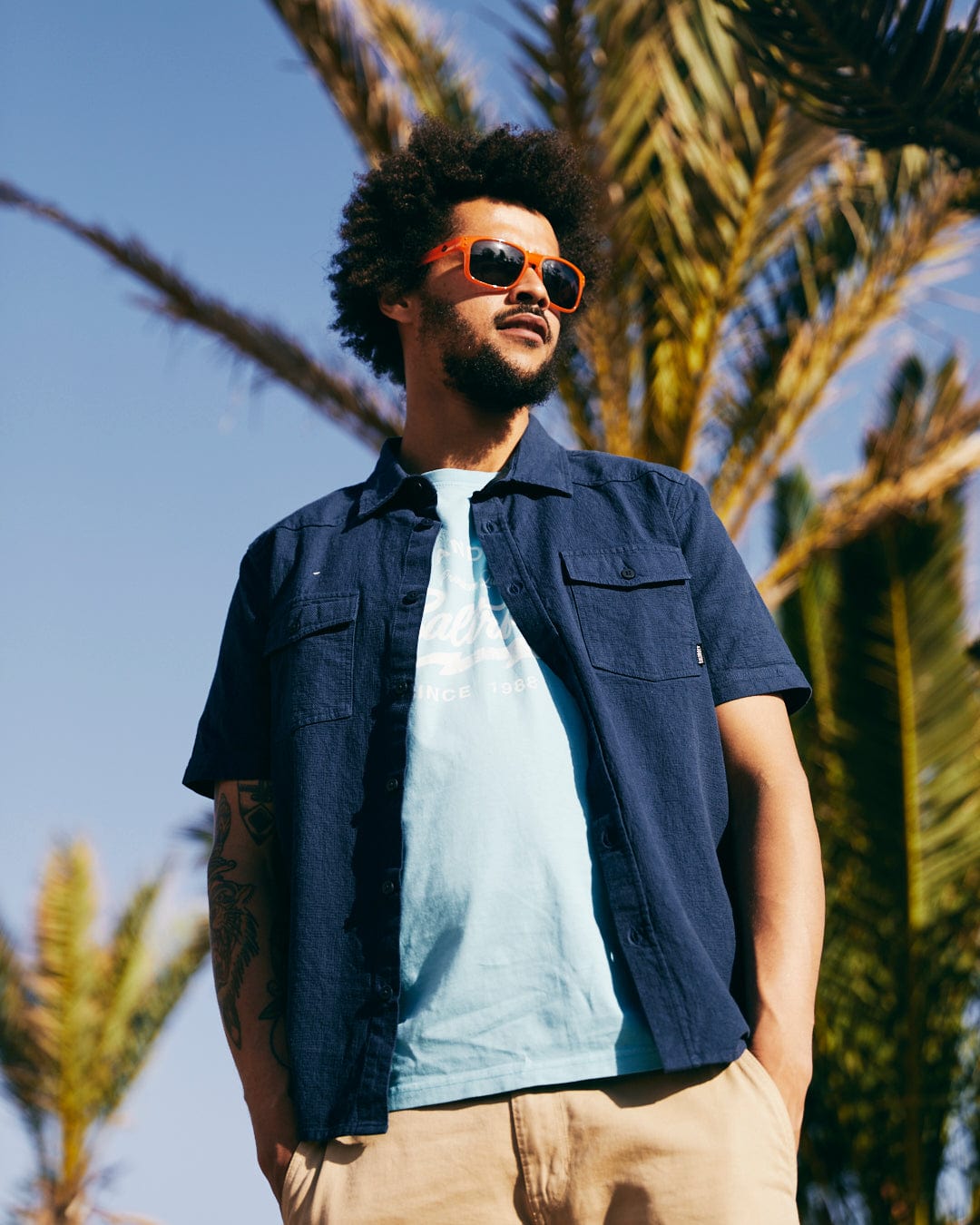 A stylish young man with an afro wearing Saltrock Ranger Recycled Polarised Sunglasses in Orange, a blue shirt, and khaki shorts stands confidently outdoors with palm trees in the background.