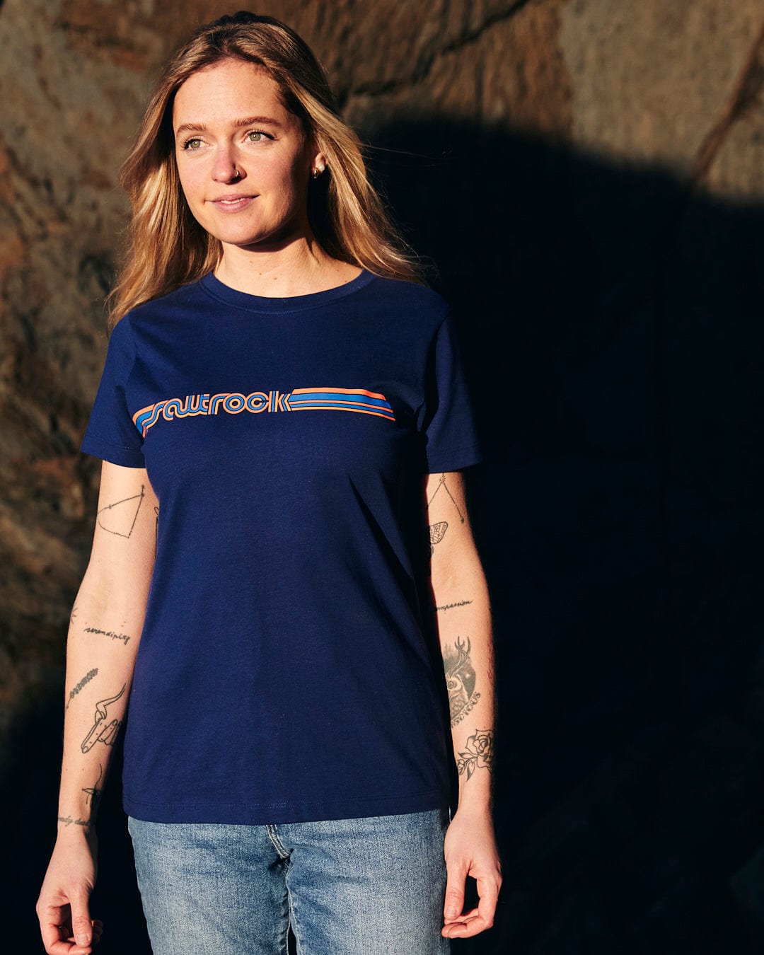 A woman wearing a Saltrock Retro Ribbon - Womens Short Sleeve T-Shirt - Dark Blue, a blue t - shirt with retro stripes, standing in front of a rock.
