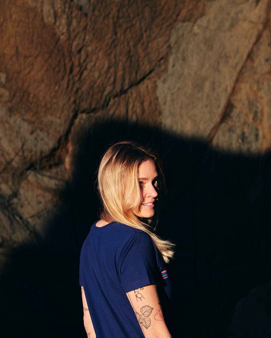 A woman in a blue Saltrock Retro Ribbon - Womens Short Sleeve T-Shirt - Dark Blue standing in front of a rock.