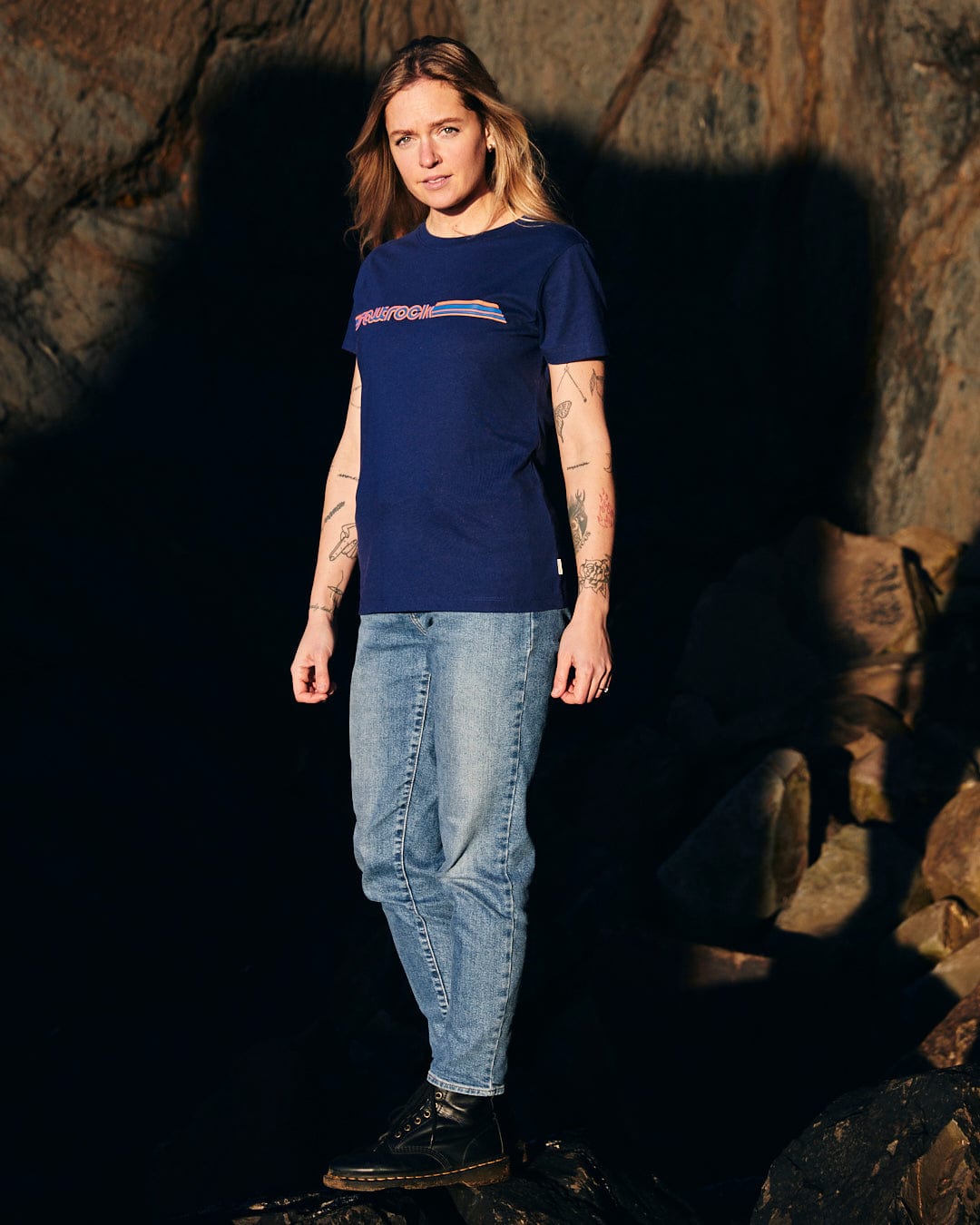 A woman wearing a Saltrock Retro Ribbon - Womens Short Sleeve T-Shirt - Dark Blue - a blue t-shirt with retro stripes - standing on rocks.