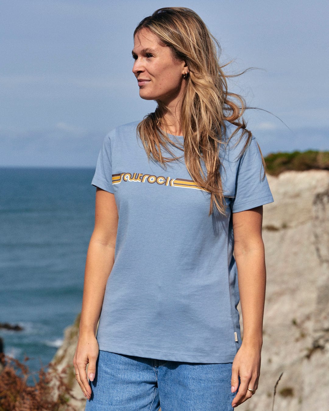 A woman wearing a Retro Ribbon - Womens Short Sleeve T-Shirt - Light Blue from the Saltrock branding stands next to the ocean.