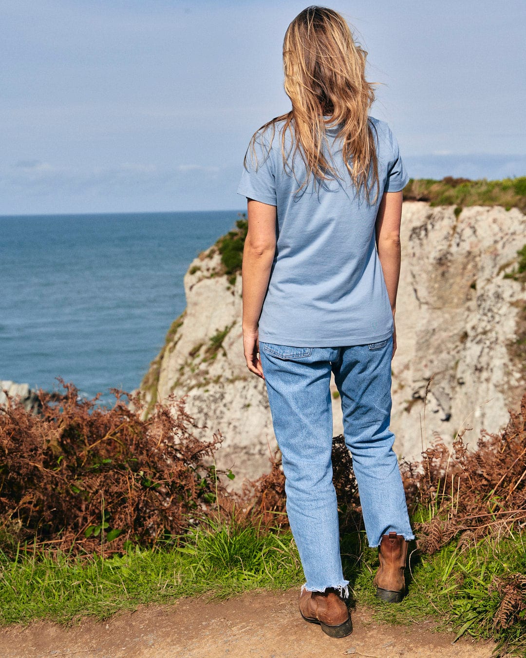 A woman is dressed in a Saltrock Retro Ribbon - Womens Short Sleeve T-Shirt - Light Blue, standing on a cliff and admiring the ocean.