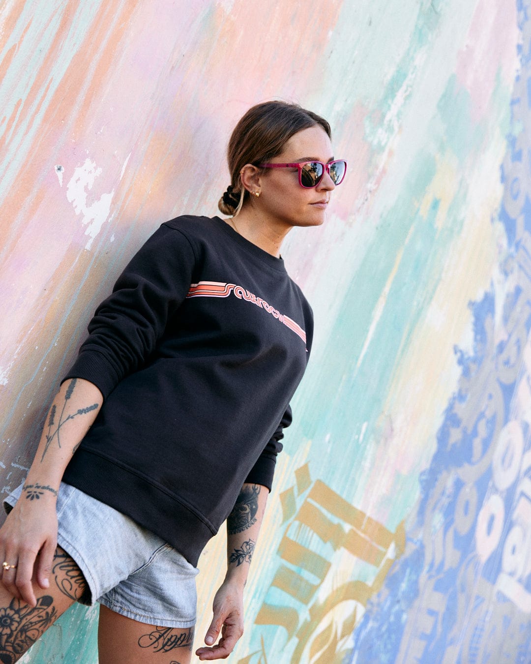 A woman in sunglasses and a Saltrock Retro Ribbon Tape - Womens Sweatshirt - Dark Grey stands in front of a colorful graffitied wall, her hands partially in her shorts pockets.