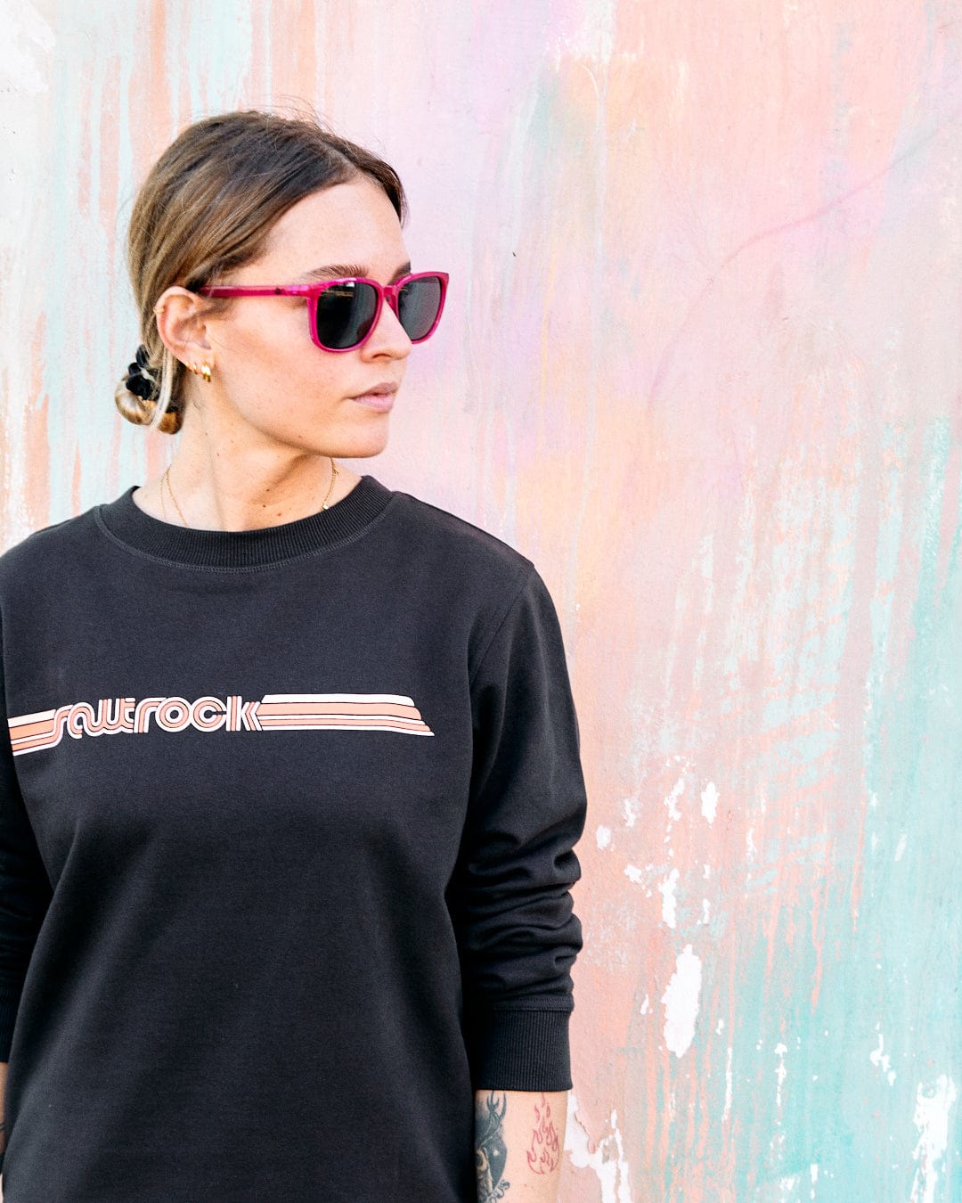 Woman in pink sunglasses and a Retro Ribbon Tape - Womens Sweatshirt - Dark Grey made of peached soft material, standing in front of a pastel-colored wall.