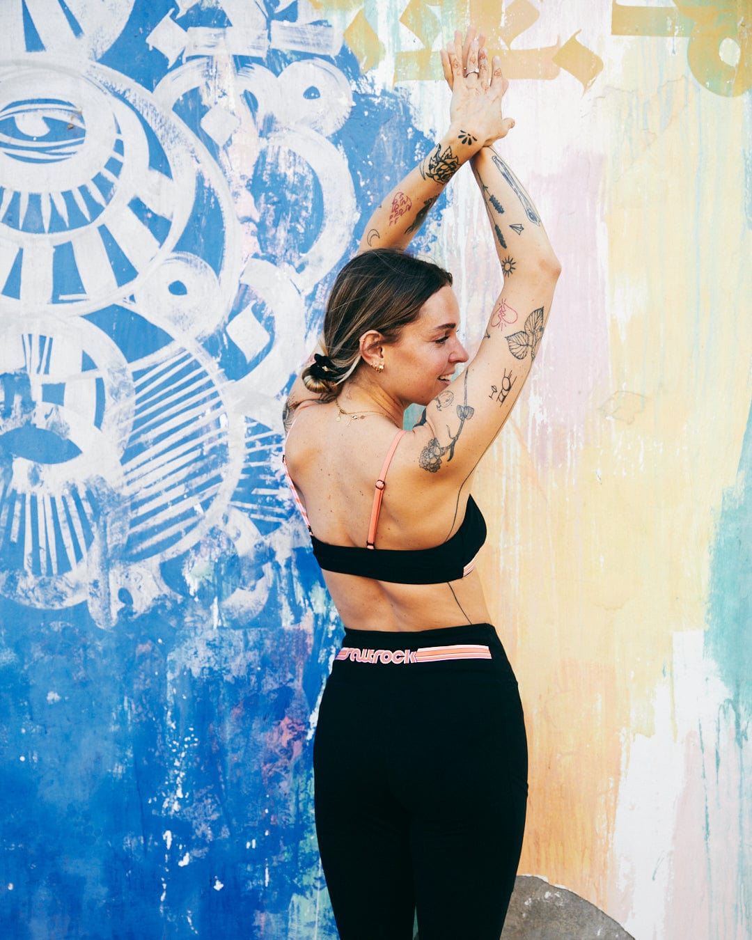 A woman with tattoos smiles and stretches her arms up against a colorful graffiti wall, wearing Retro Ribbon leggings by Saltrock, crafted from super soft fabric.