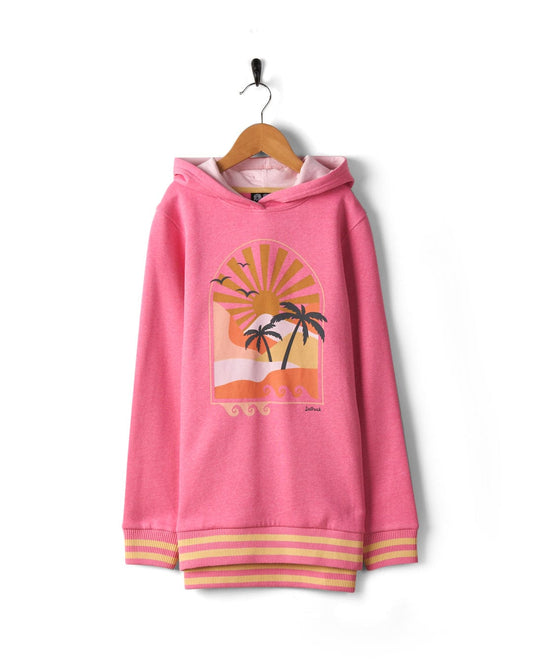 A Saltrock Retro Seascape - Recycled Kids Longline Pop Hoodie - Pink with a sunrise graphic and palm trees on the front, hanging on a wooden hanger against a white background. The cuffs and hem feature yellow and pink striped detailing.