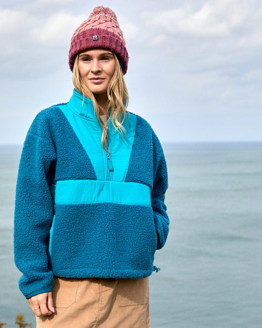 A person wearing the Saltrock Robyn Women's Fleece in blue, light brown pants, and a pink knit hat stands outdoors with the ocean and a cloudy sky in the background.