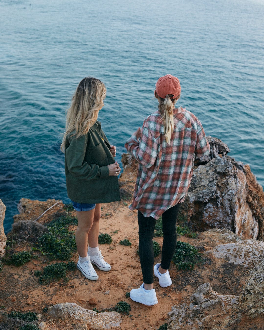 Two individuals wearing Rosalin - Womens Long Sleeve Shirts in Orange/green by Saltrock, with contrasting buttons, are standing on a rocky cliff overlooking the sea.