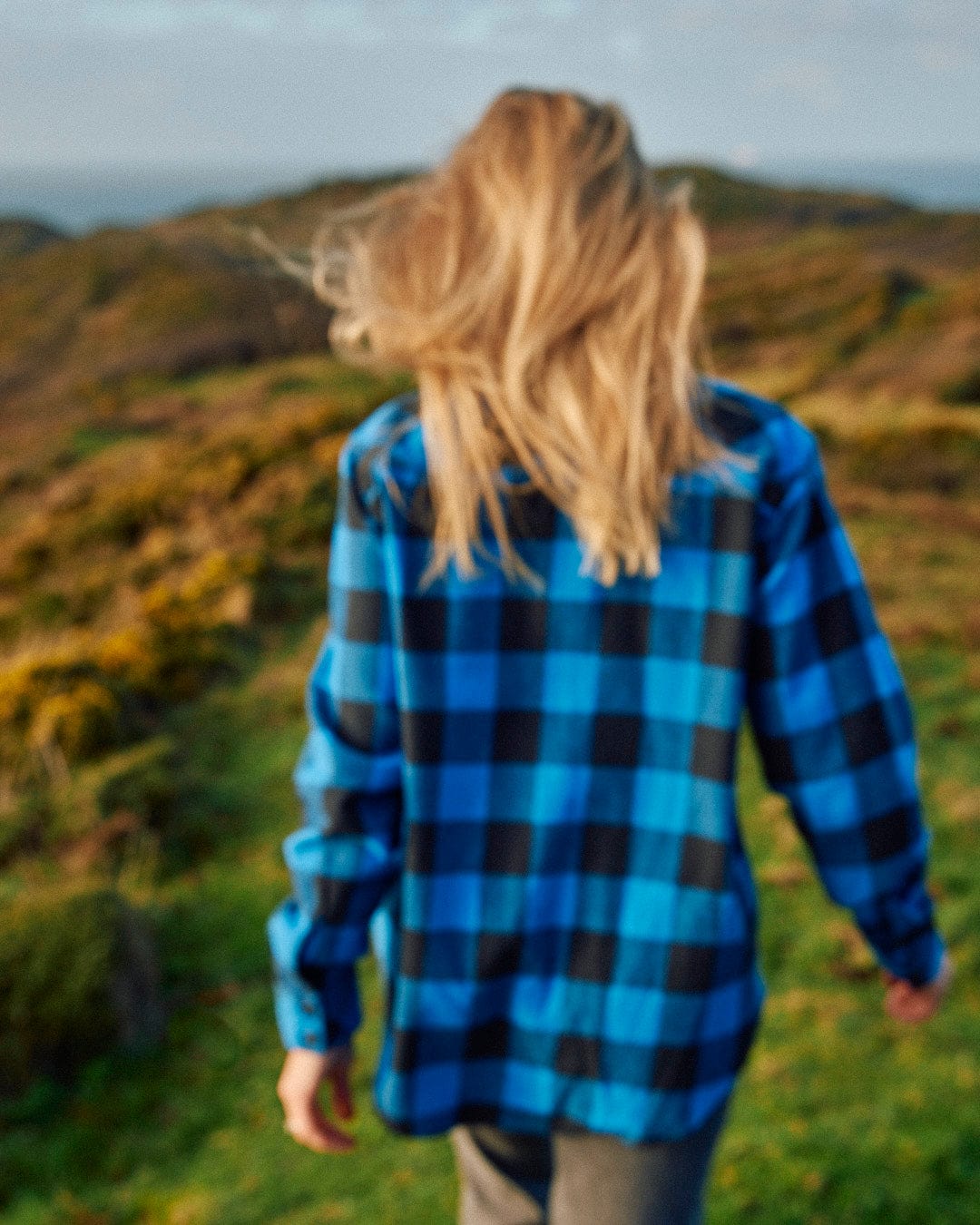 Saltrock's Rosalin - Womens Check Shirt - Blue/Black, a woman wearing a blue and black check pattern plaid shirt, walking on a grassy hill.