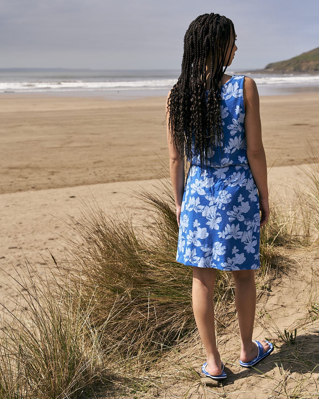 A woman in a Saltrock Floral - Womens Tie Vest Dress - Blue standing on a beach.