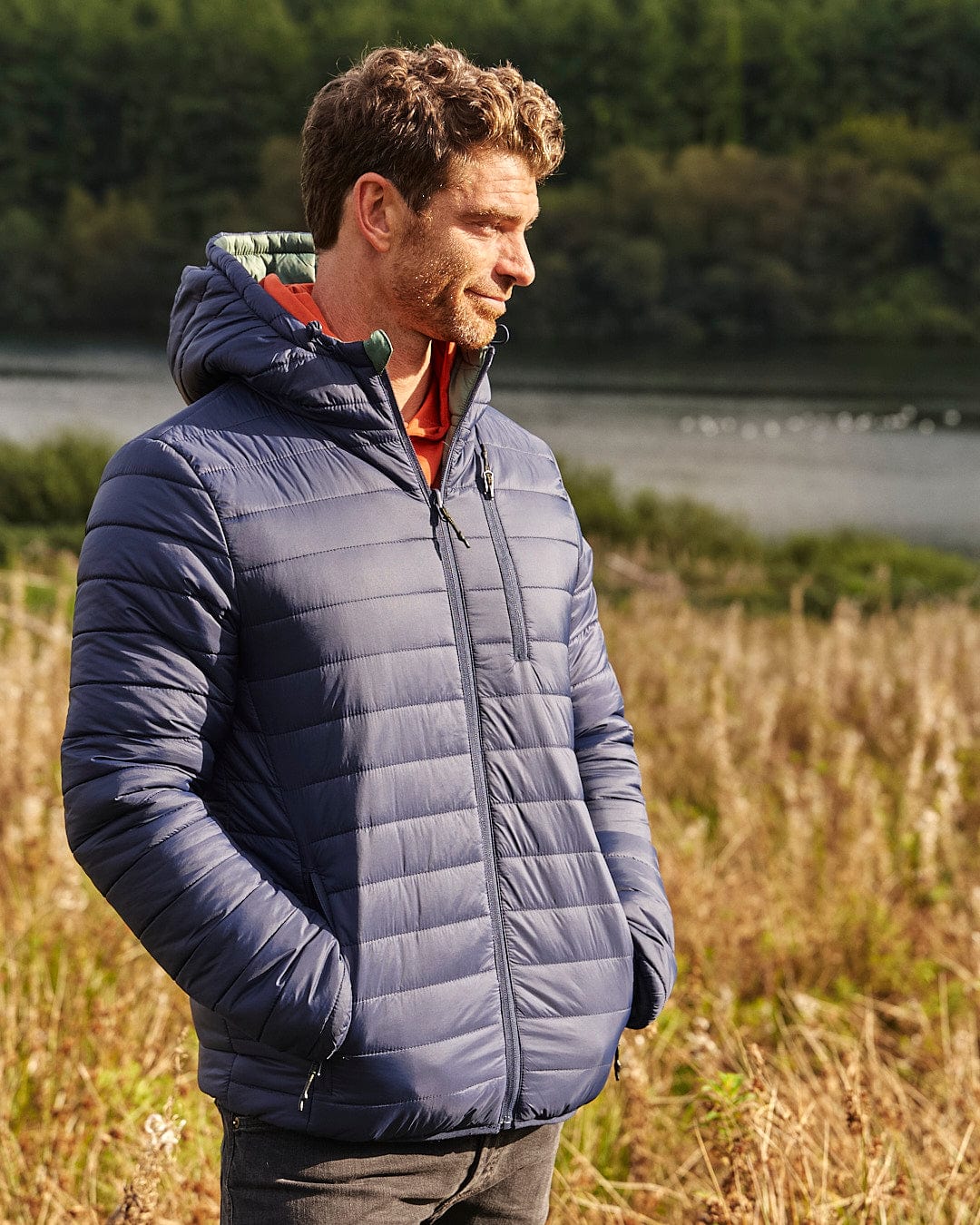 A man, dressed in a Rusik 2 - Reversible Padded Jacket by Saltrock featuring zip pockets for convenience, is standing in a water-resistant field near a lake.