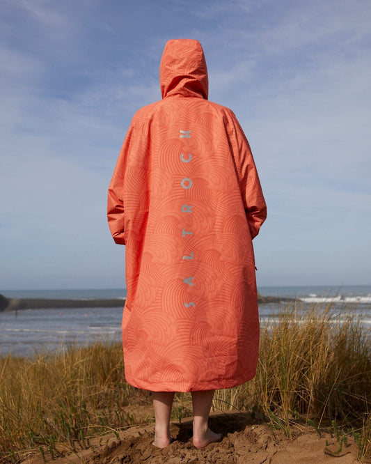 A person standing on a sandy beach wearing a Saltrock Four Seasons Changing Robe - Light Orange, in orange with the word "slowtide" patterned on the back, facing the ocean.