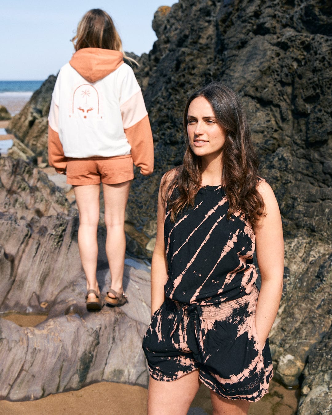 Two women are on a rocky beach; one faces the camera wearing a Saltrock Saffron - Womens Tie Front Vest - Black, while the other stands with back turned, in a white and orange hoodie with shorts. The lively attire is as practical as it is colorful, being both comfortable and machine washable.