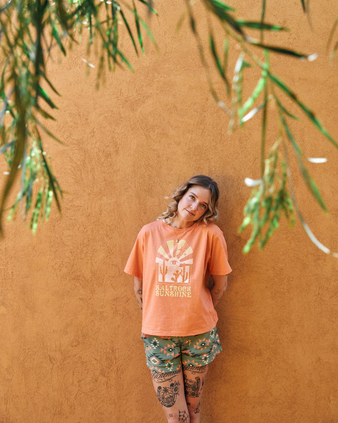 A person leans against an orange wall, wearing an orange t-shirt with Saltrock branding and patterned shorts. Featuring a boxy design with relaxed drop shoulders, the outfit blends seamlessly into the mystic desert sunset backdrop. Green foliage hangs in the foreground.
