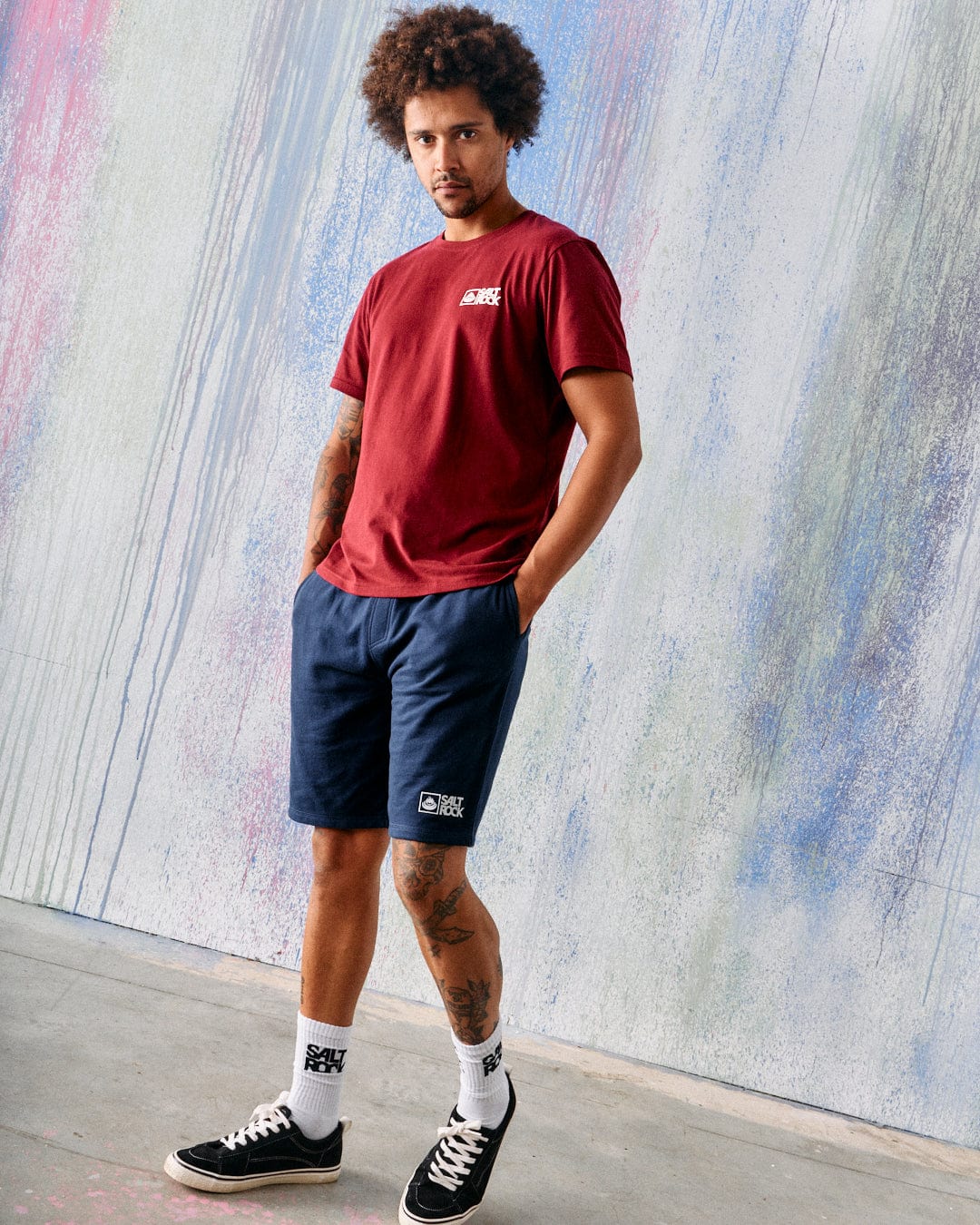A man stands indoors against a textured wall, wearing a maroon jersey T-shirt, Saltrock Original Men's Shorts in Blue Marl by Saltrock, white socks, and black sneakers.