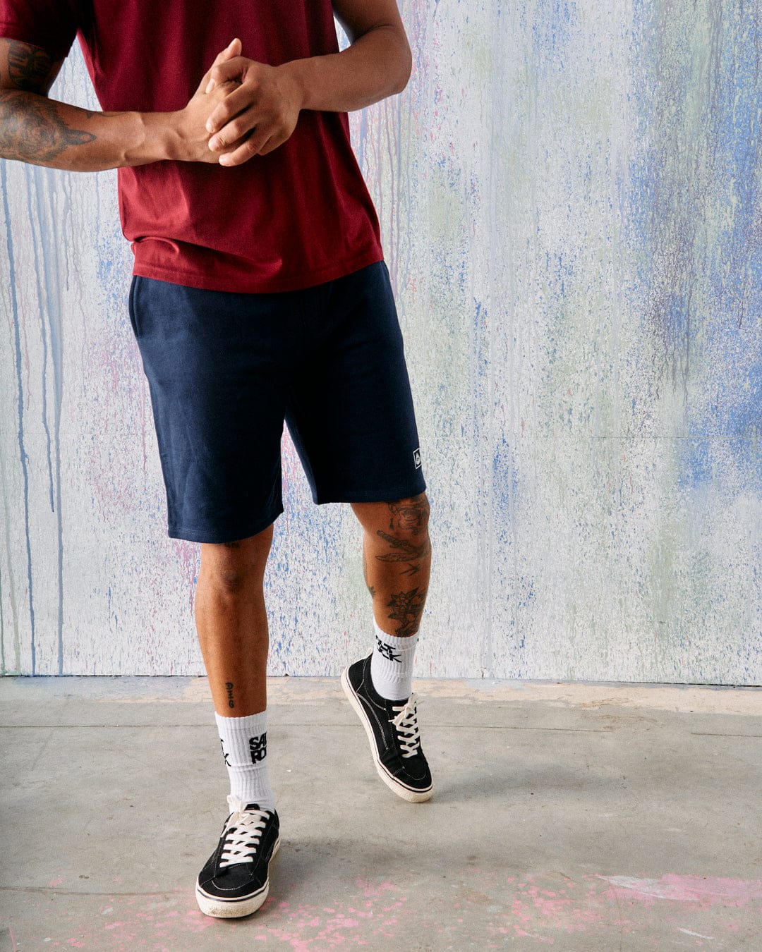 A person wearing a red Saltrock branded jersey shirt, Saltrock Original Mens Short in Blue Marl, white socks, and black sneakers stands on a concrete floor before a textured pastel wall.