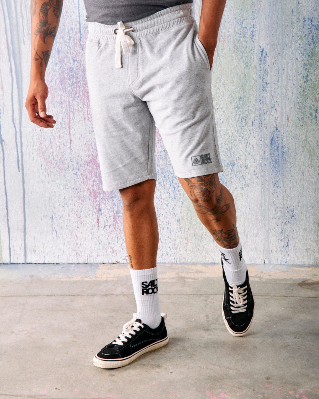 A person wears Saltrock Original Men's Shorts in grey, featuring an elasticated waist and drawstring, paired with white crew socks and black sneakers, standing on a concrete floor against a pastel textured background.