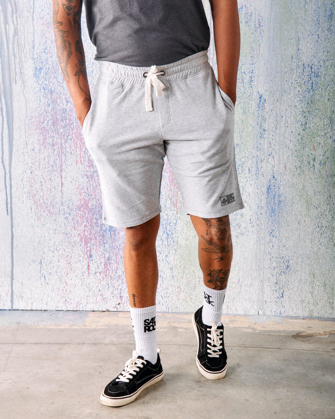 A man wears a gray t-shirt, Saltrock Original - Mens Short - Grey by Saltrock, white socks, and black sneakers, standing indoors against a textured wall with hands in his pockets.