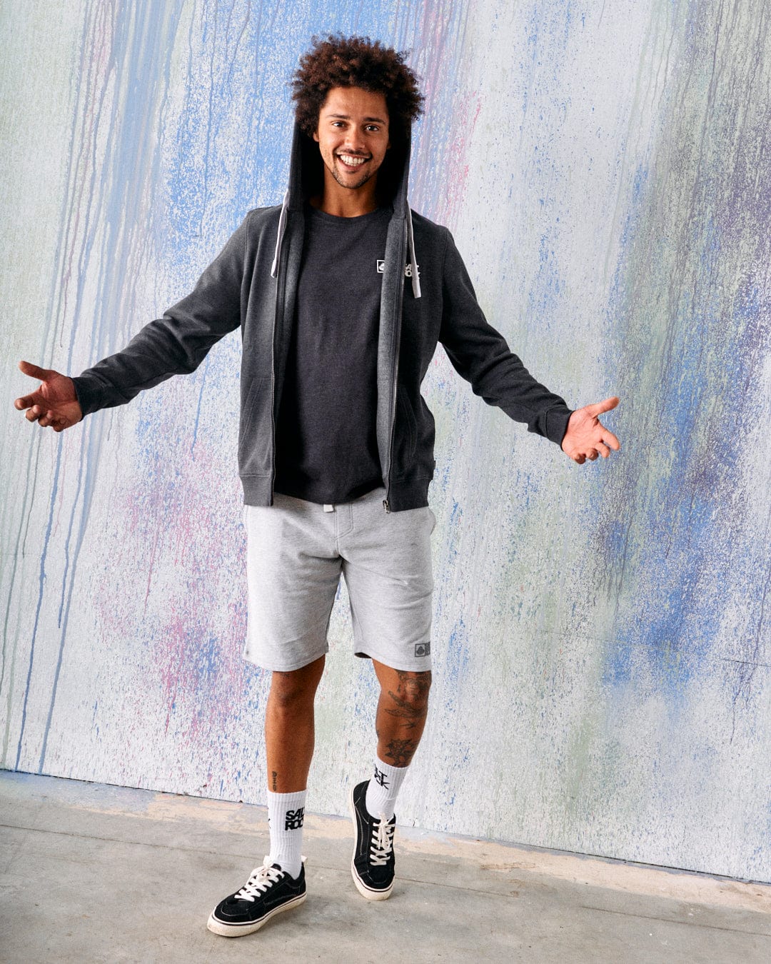 A man stands indoors, smiling with his arms open in front of a colorful textured wall, wearing a dark hoodie, black t-shirt, and Saltrock Original - Mens Short - Grey from Saltrock.