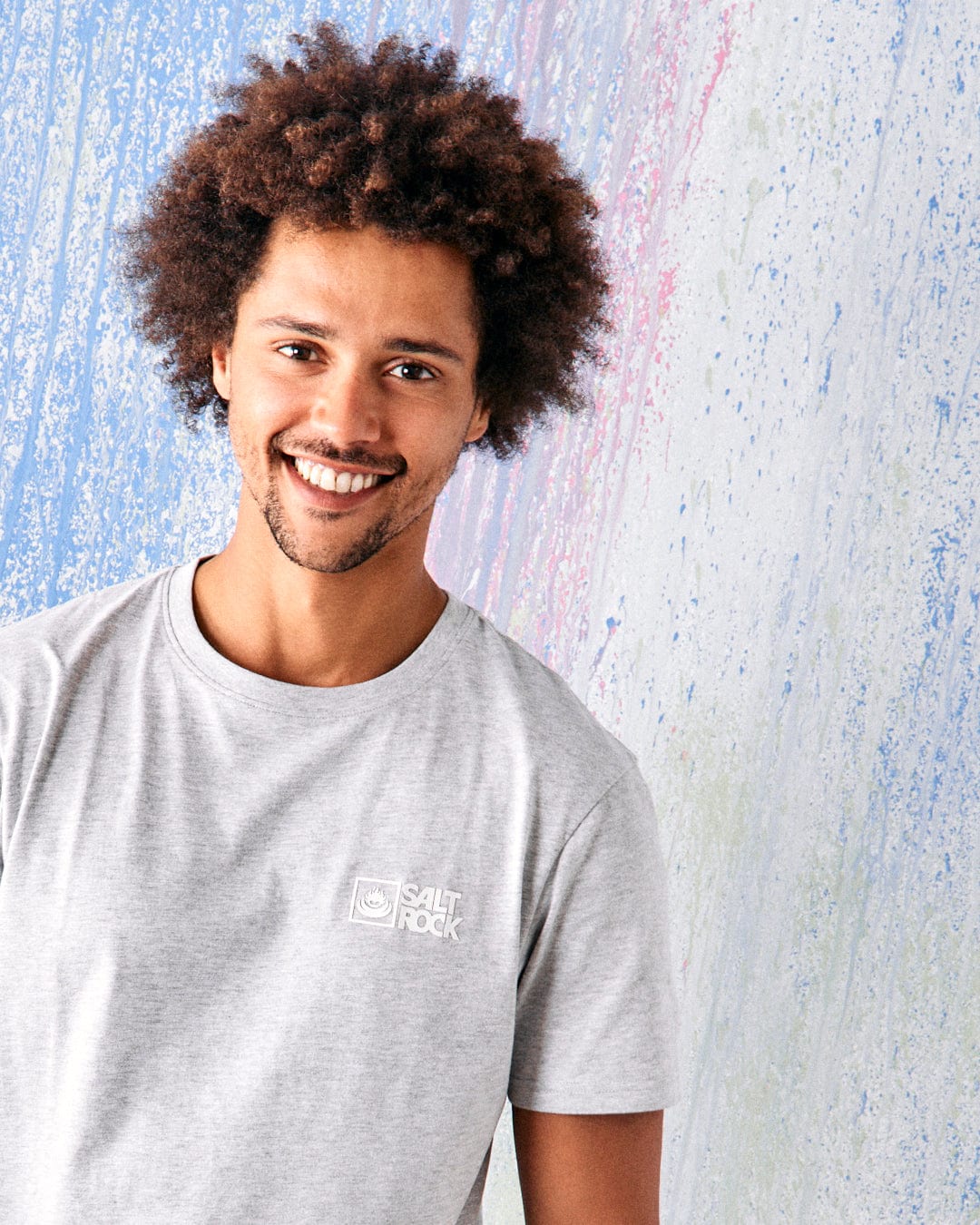 A smiling man with curly hair wears the Saltrock Original Men's Short Sleeve T-Shirt in grey, standing before a pastel textured wall.