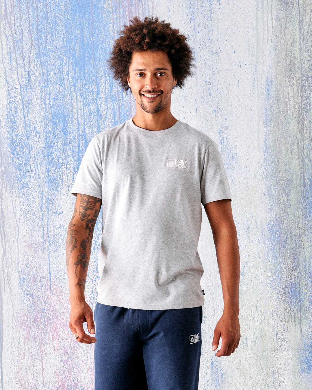 A man with curly hair and tattoos on his left arm wears the Saltrock Original Men's Short Sleeve T-Shirt in grey, standing in front of a textured pastel background.