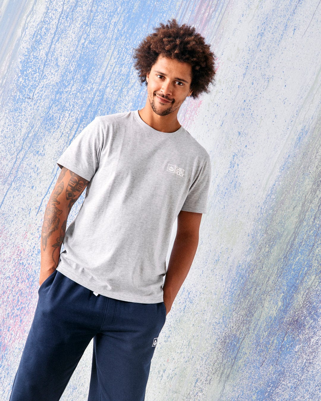 A man with curly hair wears the Saltrock Original Men's Short Sleeve T-Shirt in grey by Saltrock, paired with navy blue pants, standing against a pastel-colored textured background.