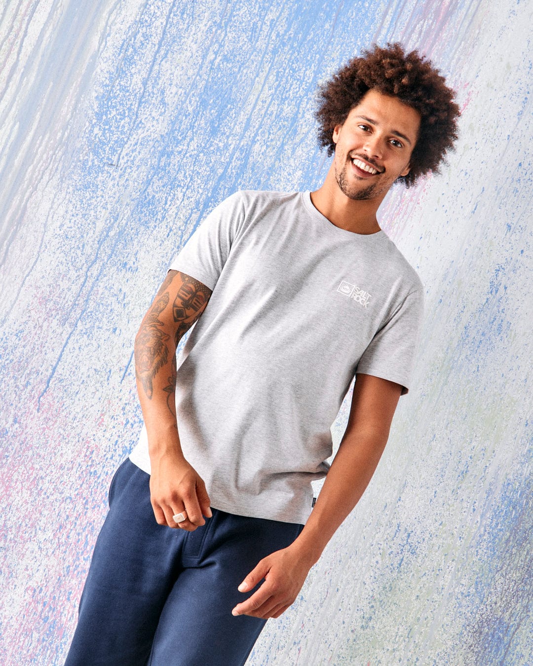 A man with curly hair and a left arm tattoo wears the Saltrock Original Mens Short Sleeve T-Shirt in grey, standing before a pastel splatter-patterned background.