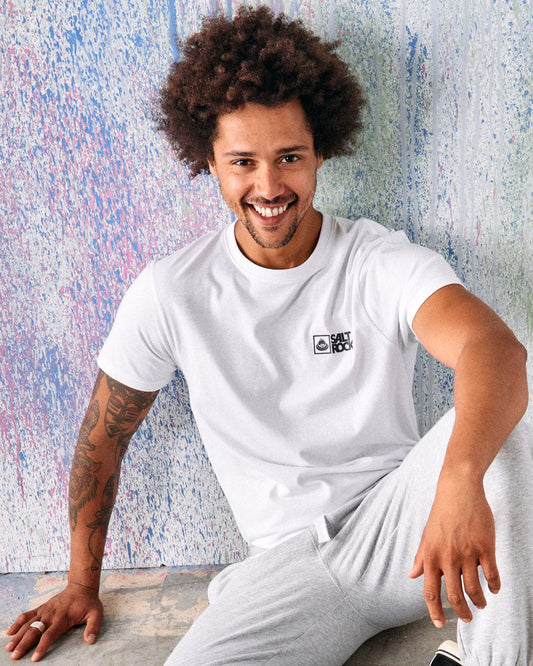 A man with curly hair and tattoos, wearing the Saltrock Original Men's Short Sleeve T-Shirt in white and light gray pants, sits on the floor smiling in front of a colorful, textured wall.