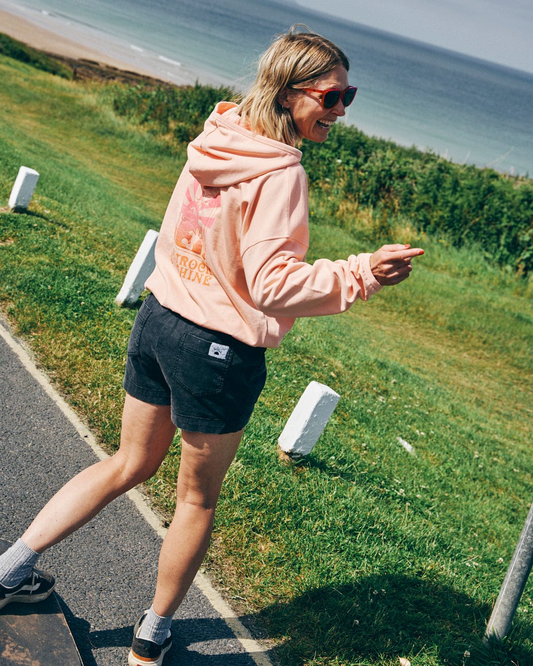 A person in a Saltrock Sunshine Pop Hood in peach and black shorts smiles while pointing, with the seaside in the background.
