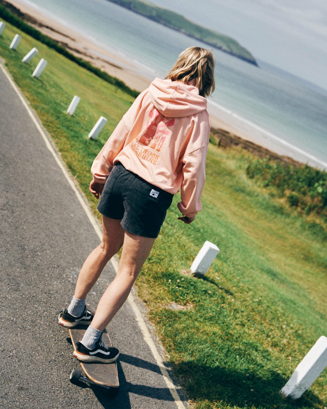 A person skateboards on a paved path near a beach, surrounded by grassy areas, wearing a peach-colored Saltrock Sunshine women's pop hoodie and black shorts, embracing the relaxed fit that complements their effortless glide.