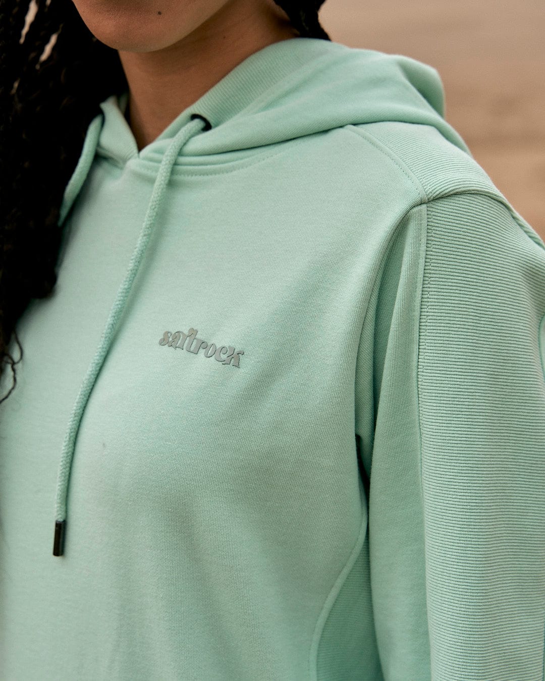A woman in a Shelley - Womens Pop Hoodie - Light Green by Saltrock is standing on the beach.