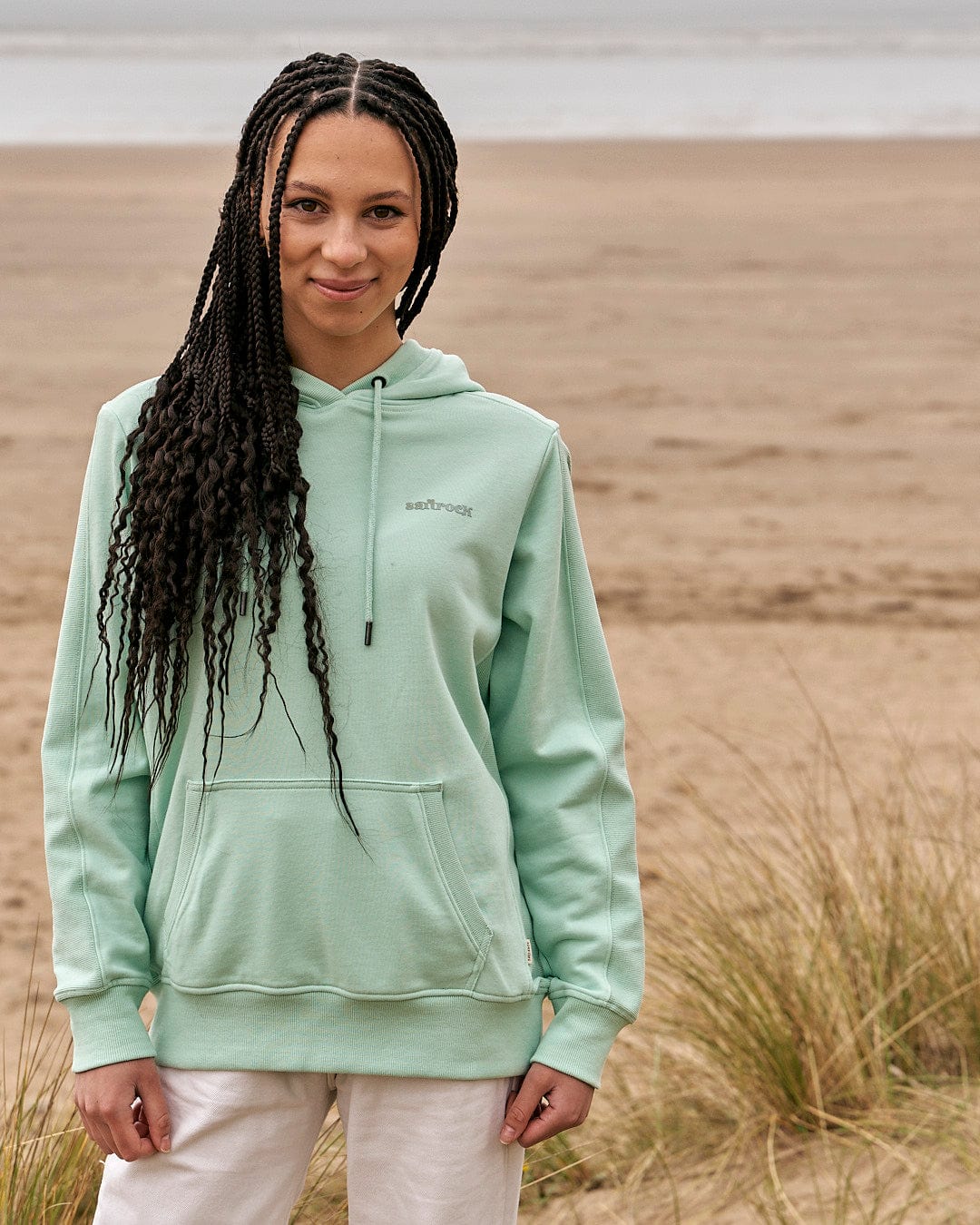 A woman wearing a Shelley - Womens Pop Hoodie - Light Green by Saltrock standing on the beach.