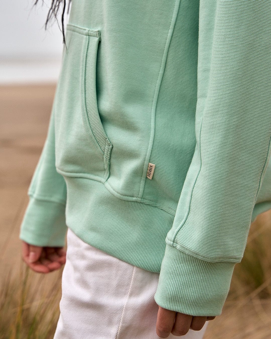 A woman wearing a Saltrock Shelley - Womens Pop Hoodie in Light Green on a beach.