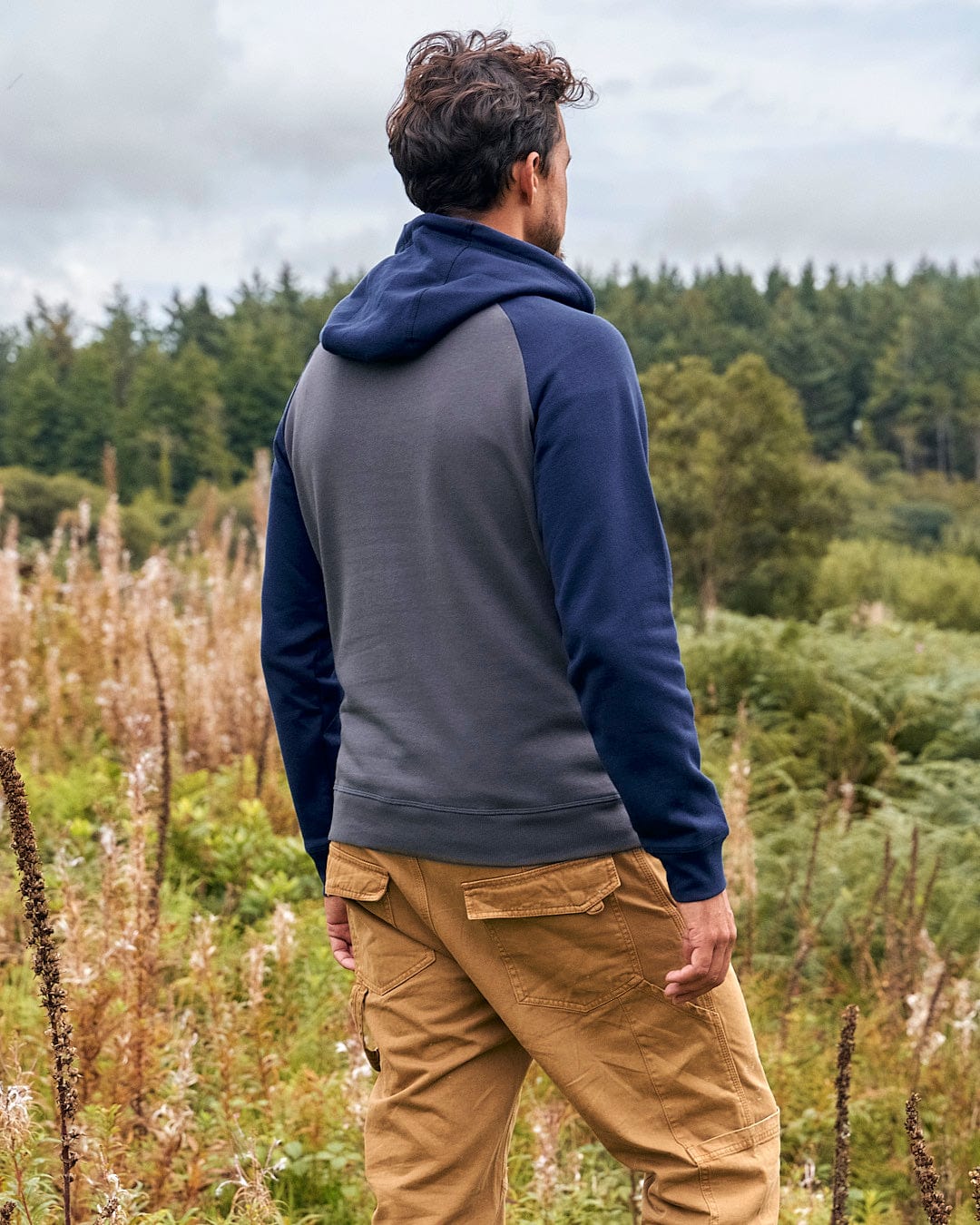 A man in a Saltrock Skelly Rider - Mens Pop Hoodie - Dark Grey stands in a field, wearing khaki pants.