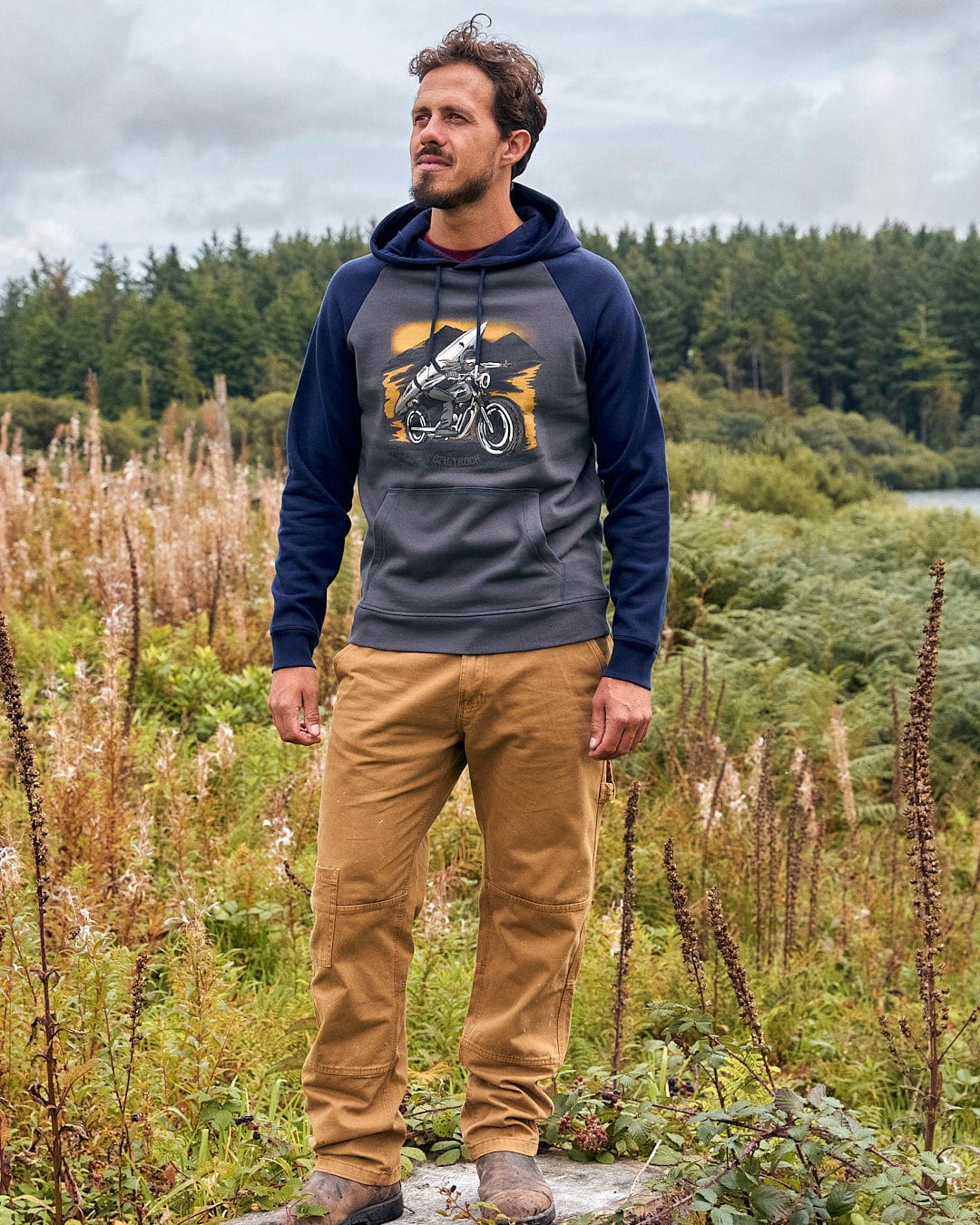 A man wearing a Saltrock Skelly Rider - Mens Pop Hoodie - Dark Grey posing in a field.