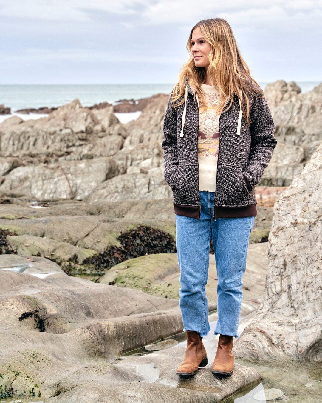 A woman wearing a Saltrock badge stands on textured knit rocks next to the ocean, donning a Sofie - Womens Borg Lined Zip Hoodie - Brown.