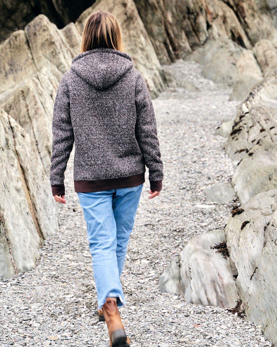 A woman wearing a textured knit Saltrock Sofie Hoodie, walking on a gravel path near rocks.