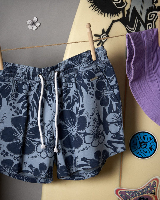 A pair of Soifra Hibiscus - Womens Sweat Short - Blue with an elasticated waist, hanging on a hanger next to a surfboard.